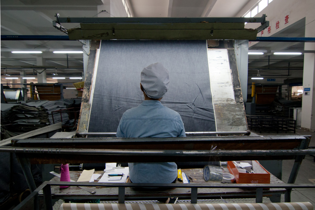 Sitting in front of a large machine that spools denim at a high speed, a Chinese factory worker examines the fabric as it spins by.