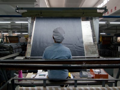 Sitting in front of a large machine that spools denim at a high speed, a Chinese factory worker examines the fabric as it spins by.