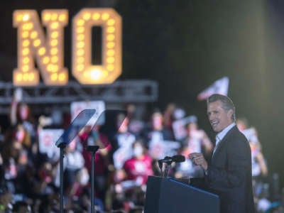 Gavin Newsom speaks in front of a huge sign reading "NO"