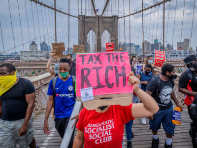 A protester in a red DEMOCRATIC SOCIALIST CLUB t-shirt holds a sign reading "TAX THE RICH" at a protesy\t