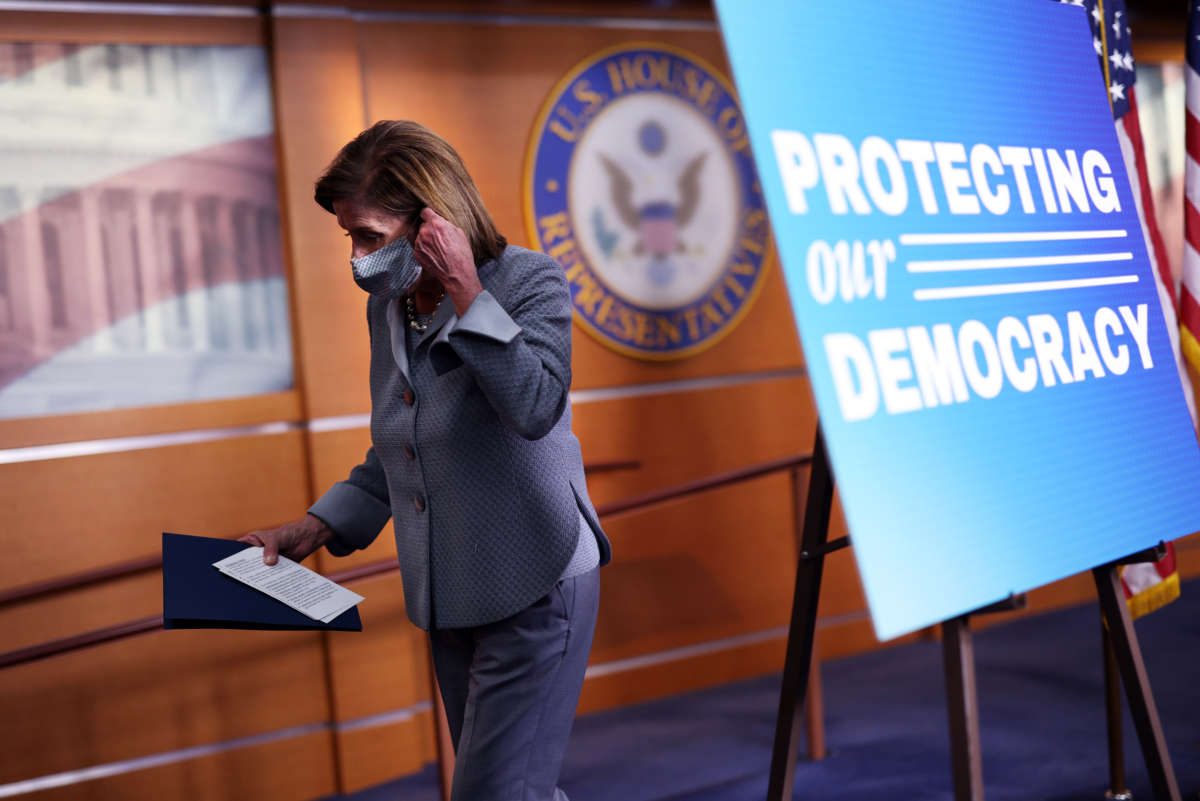 Nancy Pelosi prepares to remove her mask while walking behind a sign reading "PROTECTING OUR DEMOCRACY"