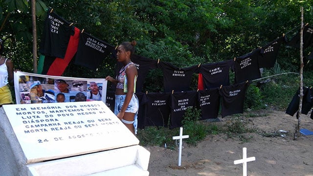 Two residents of Vila Moises stand by a banner with the pictures of the victims of the Cabula Massacre alongside black t-shirts that bear each victim's name.