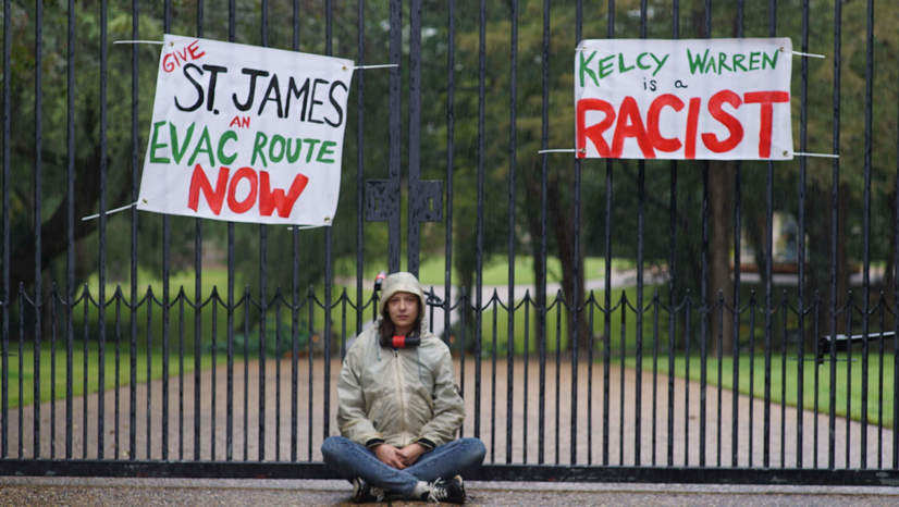 A protester sits in front of the gates to Energy Transfer CEO Kelcy Warren's Dallas home, in front of signs objecting to the lack of an evacuation route for an African American community in St. James Parish, Louisiana, the terminus of the Bayou Bridge Pipeline.