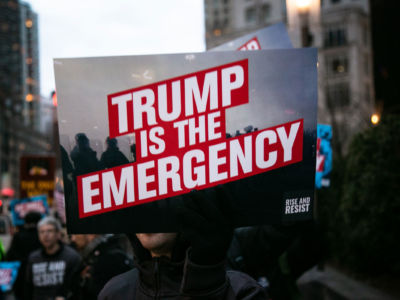 A person holds protest sign that reads "Trump is the emergency" in front of Trump International Hotel on February 15, 2019, in New York City. The group is protesting President Donald Trump's declaration of a National Emergency in order to build his proposed border wall.