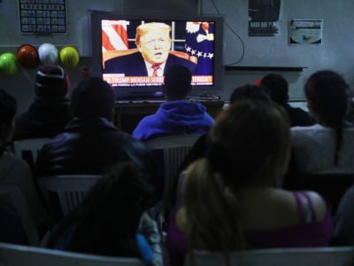 Migrants view a live televised speech by President Donald Trump on border security at a shelter for migrants on January 8, 2019, in Tijuana, Mexico.
