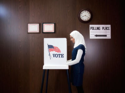 Hijabi in denim dress stands at voting booth bearing the U.S. flag