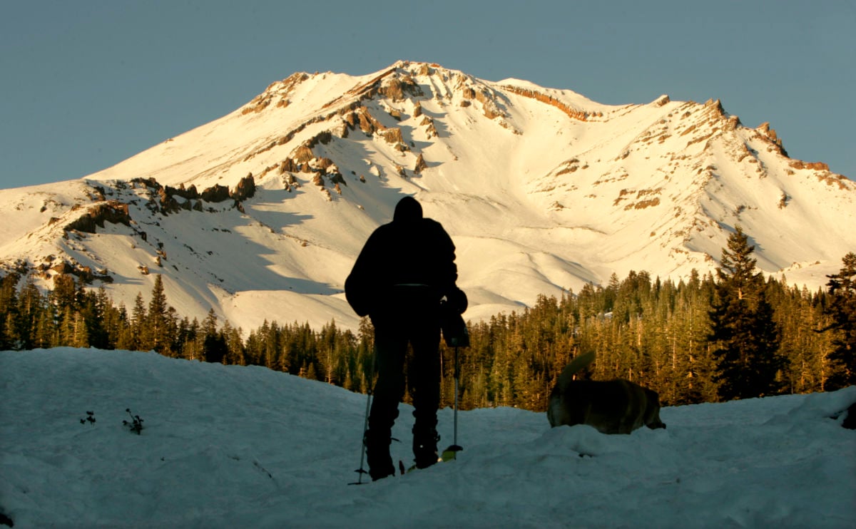 A hiker standing in the snow is silhouetted against a bright, snow-capped mountain