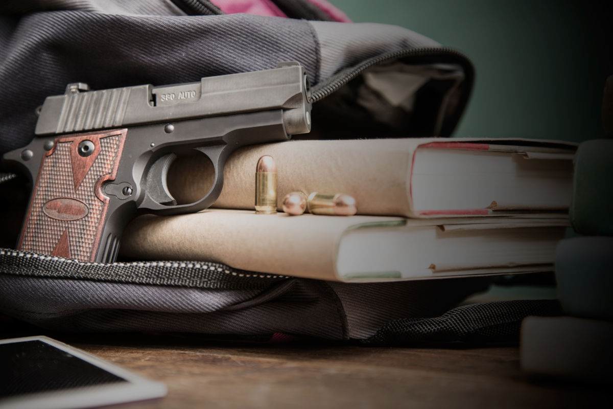 A gun sticks out of a backpack on a school desk with textbooks nearby