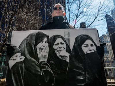 An older man holds a black and white photo of crying women in chador