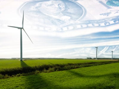 Wind turbines stand on green plains in front of a blue sky with a filter of U.S dollars