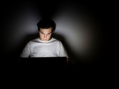 A young white man is illuminated by his laptop's screen in a darkened room