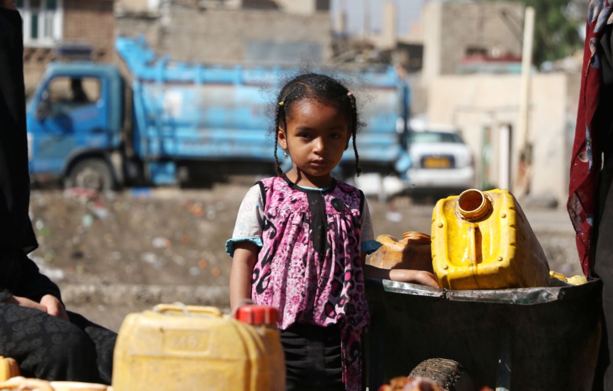 A young girl stands by two yellow water buckets