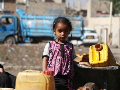 A young girl stands by two yellow water buckets