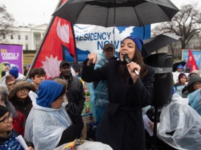 AOC speaks during an event in the rain, surrounded by supporters