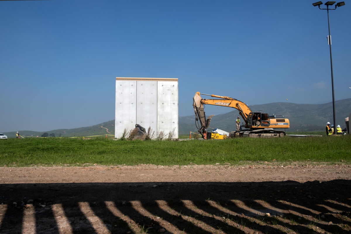 A bulldozer sits next to a solid grey wall prototype