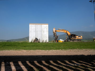 A bulldozer sits next to a solid grey wall prototype