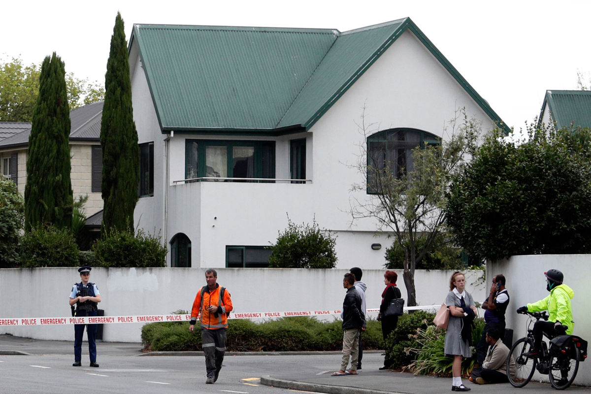 People stand in front of a white building with a green roof, cordoned off with police tape