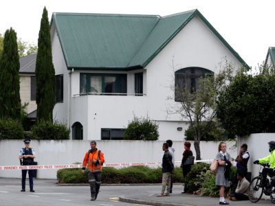 People stand in front of a white building with a green roof, cordoned off with police tape