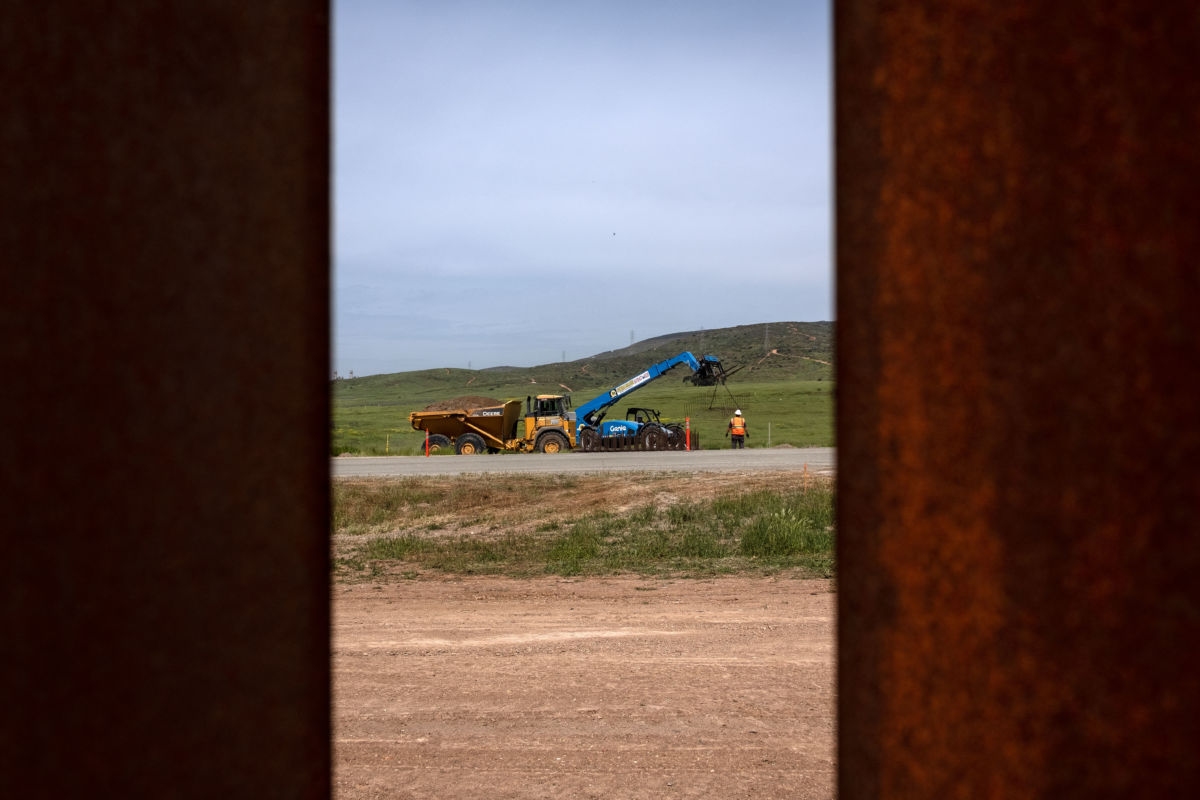 a bulldozer and workers are seen through rusted slats of the existing border fence