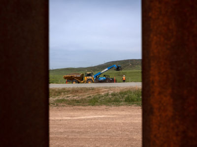 a bulldozer and workers are seen through rusted slats of the existing border fence