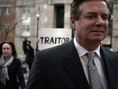 Paul Manafort walks away from man holding a sign reading "TRAITOR"
