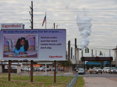 A smokestack pours out smoke behind a sign claiming the safety of Exxon Mobil products