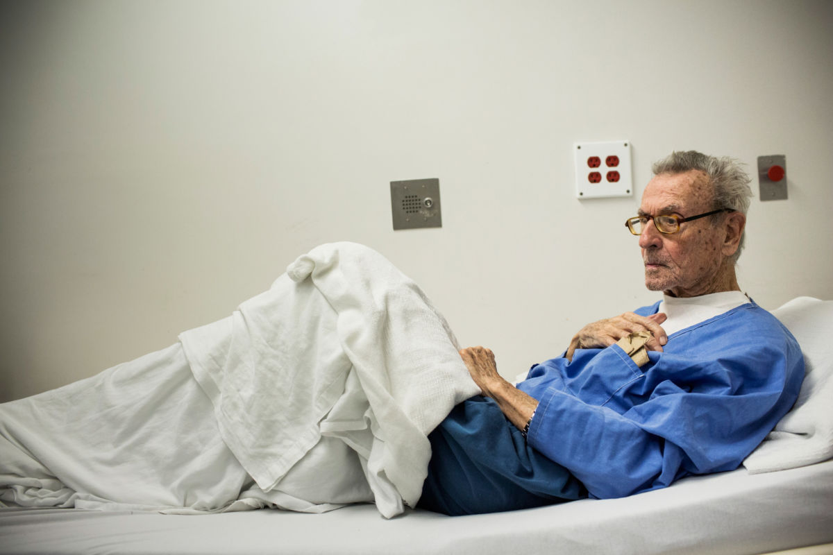 An elderly prisoner sits in a hospital bed