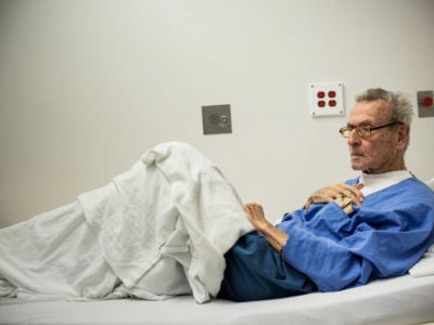 An elderly prisoner sits in a hospital bed