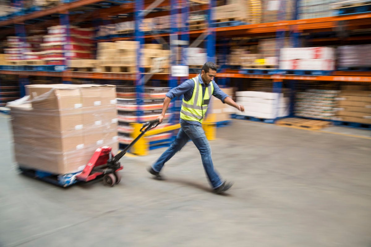A man hurries through a warehouse pulling a dolly stacked with boxes