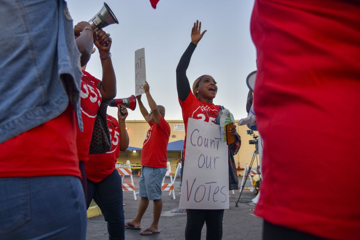 Women in red shirts demonstrate with a sign reading "COUNT OUR VOTES"