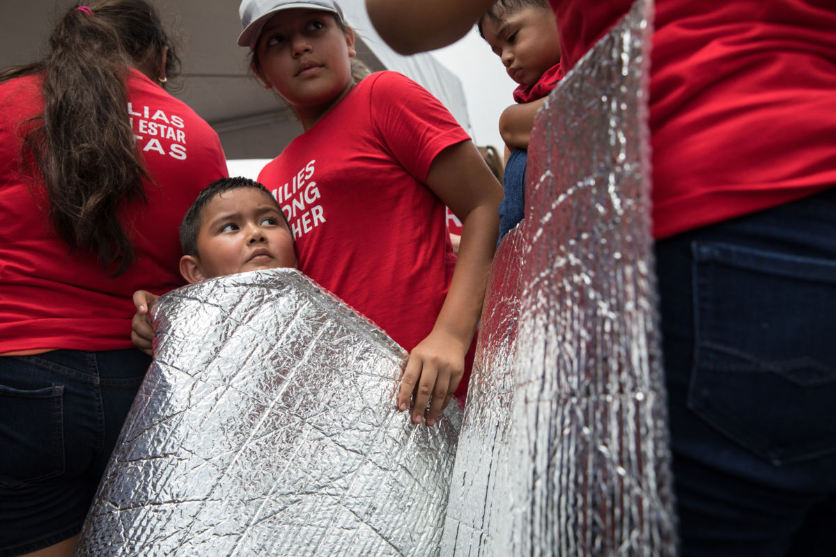 A young boy wearing the same red shirt as the adults surrounding him holds a thermal blanket