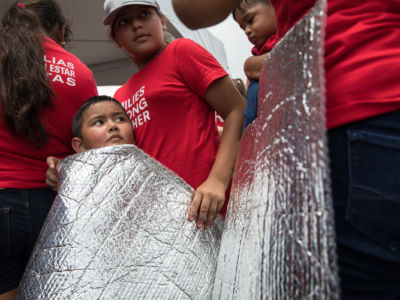 A young boy wearing the same red shirt as the adults surrounding him holds a thermal blanket