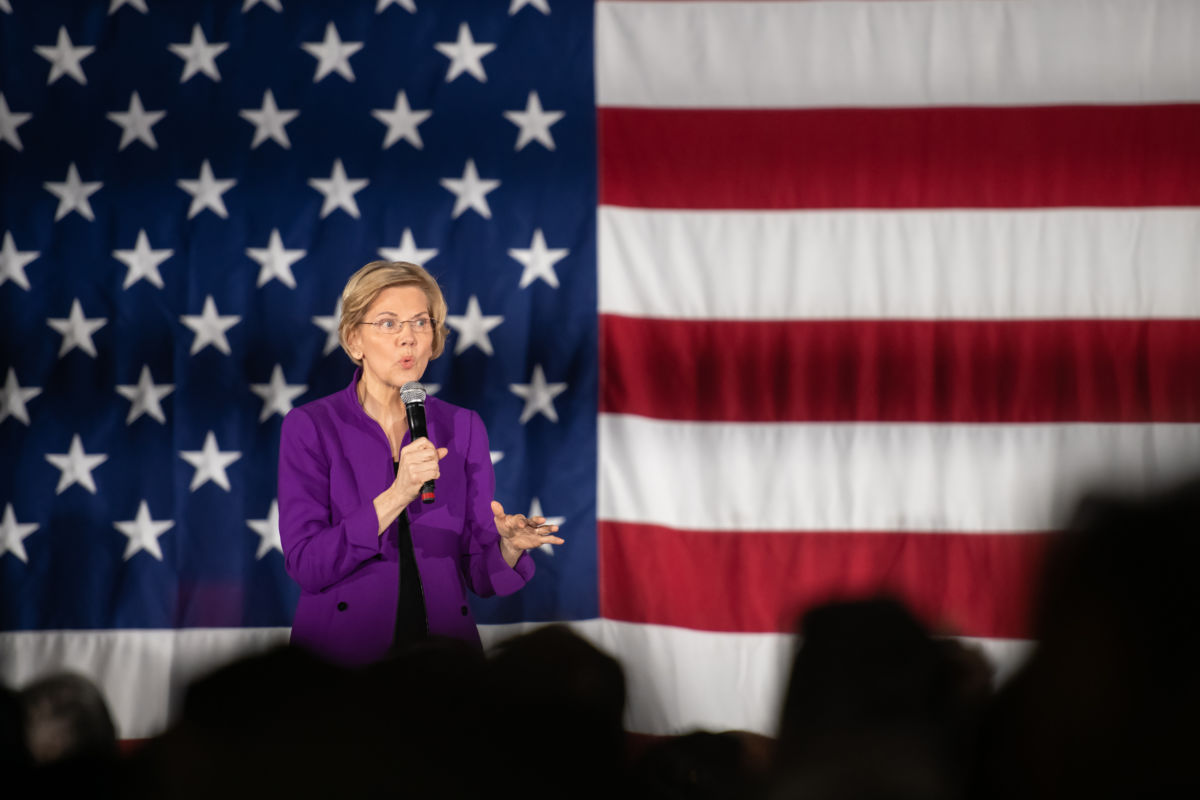 Sen. Elizabeth Warren speaks into a microphone while standing in front of a U.S Flag