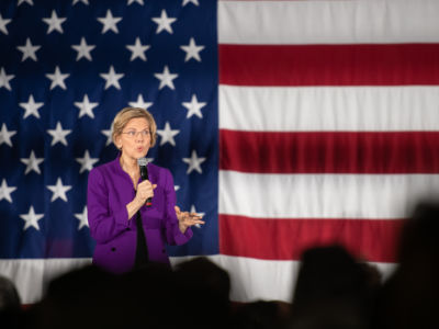 Sen. Elizabeth Warren speaks into a microphone while standing in front of a U.S Flag