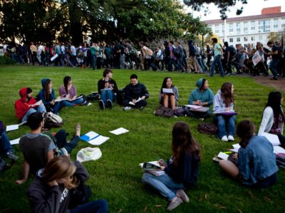 Students sit in a circle on a grassy lawn as people walk to a protest behind them