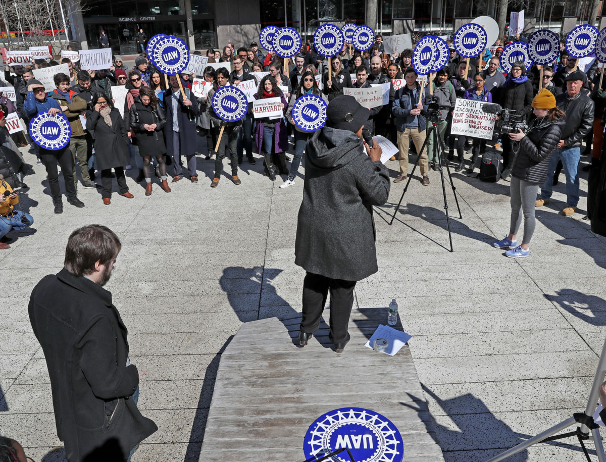 A person speaks to a crowd holding blue signs reading (UAW)