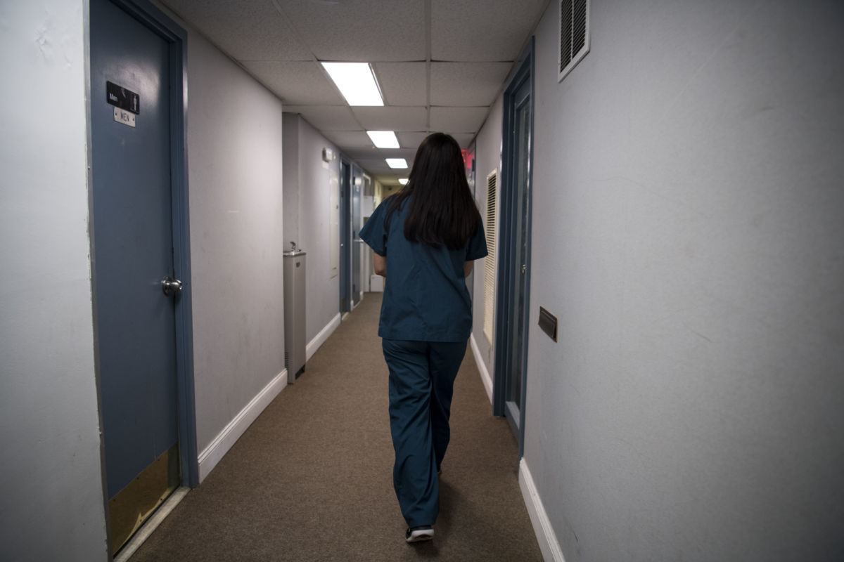 A woman in teal scrubs walks down a clinic hallway
