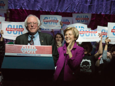 Elizabeth Warren and Bernie Sanders stand at a podium