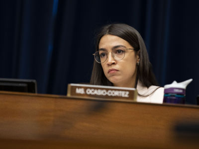 Alexandria Ocasio-Cortez listens while seated at a podium