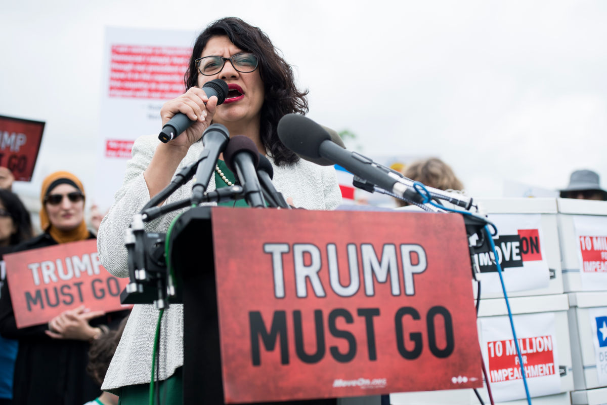 Rep. Rashida Tlaib speaks into a microphone behind a sign reading "TRUMP MUST GO"