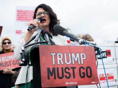 Rep. Rashida Tlaib speaks into a microphone behind a sign reading "TRUMP MUST GO"