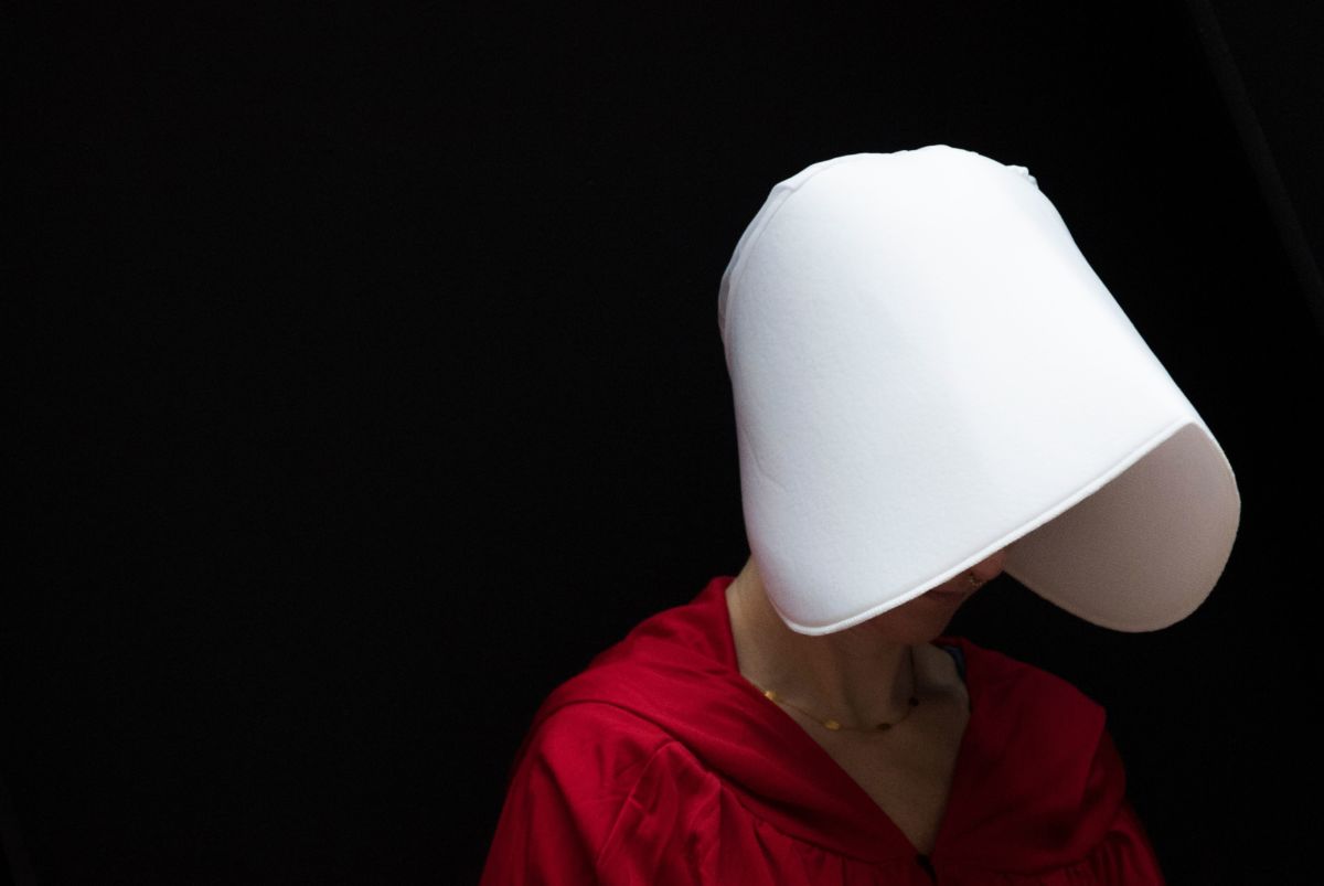 A woman's face is obscured by a white bonnet as she wears a red robe