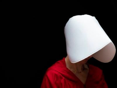 A woman's face is obscured by a white bonnet as she wears a red robe