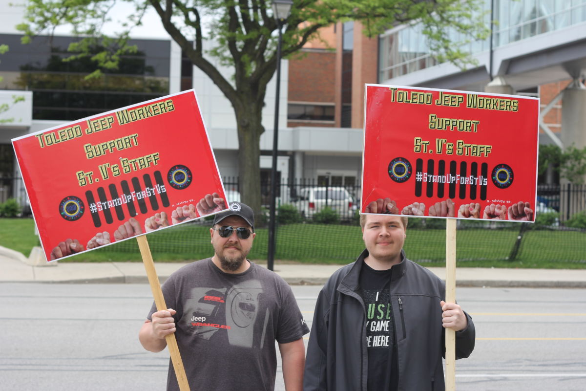 Two men display red signs reading "TOLEDO JEEP WORKERS SUPPORT ST. V'S STAFF"