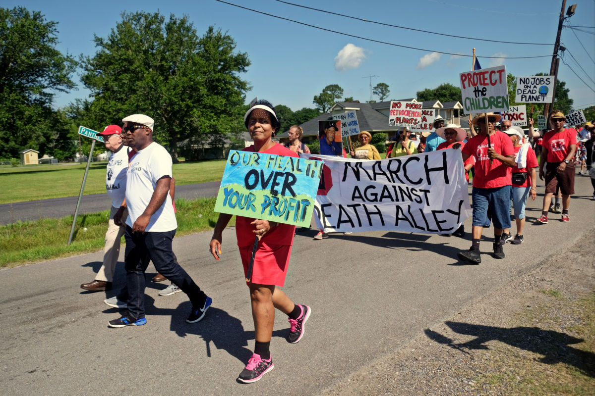 A woman wearing red protests while holding a sign that reads "OUR HEALTH OVER YOUR PROFITS"