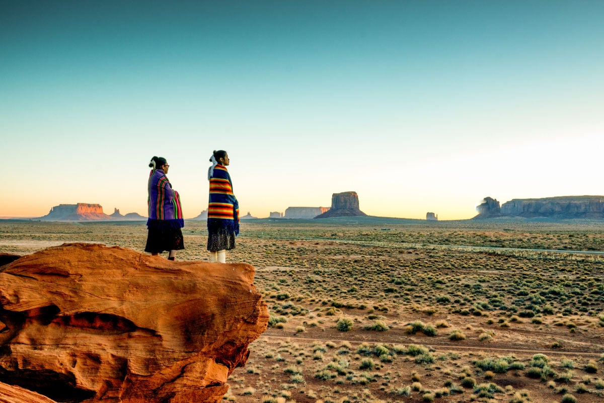Two Navajo women stand on a cliff overlooking rock formations