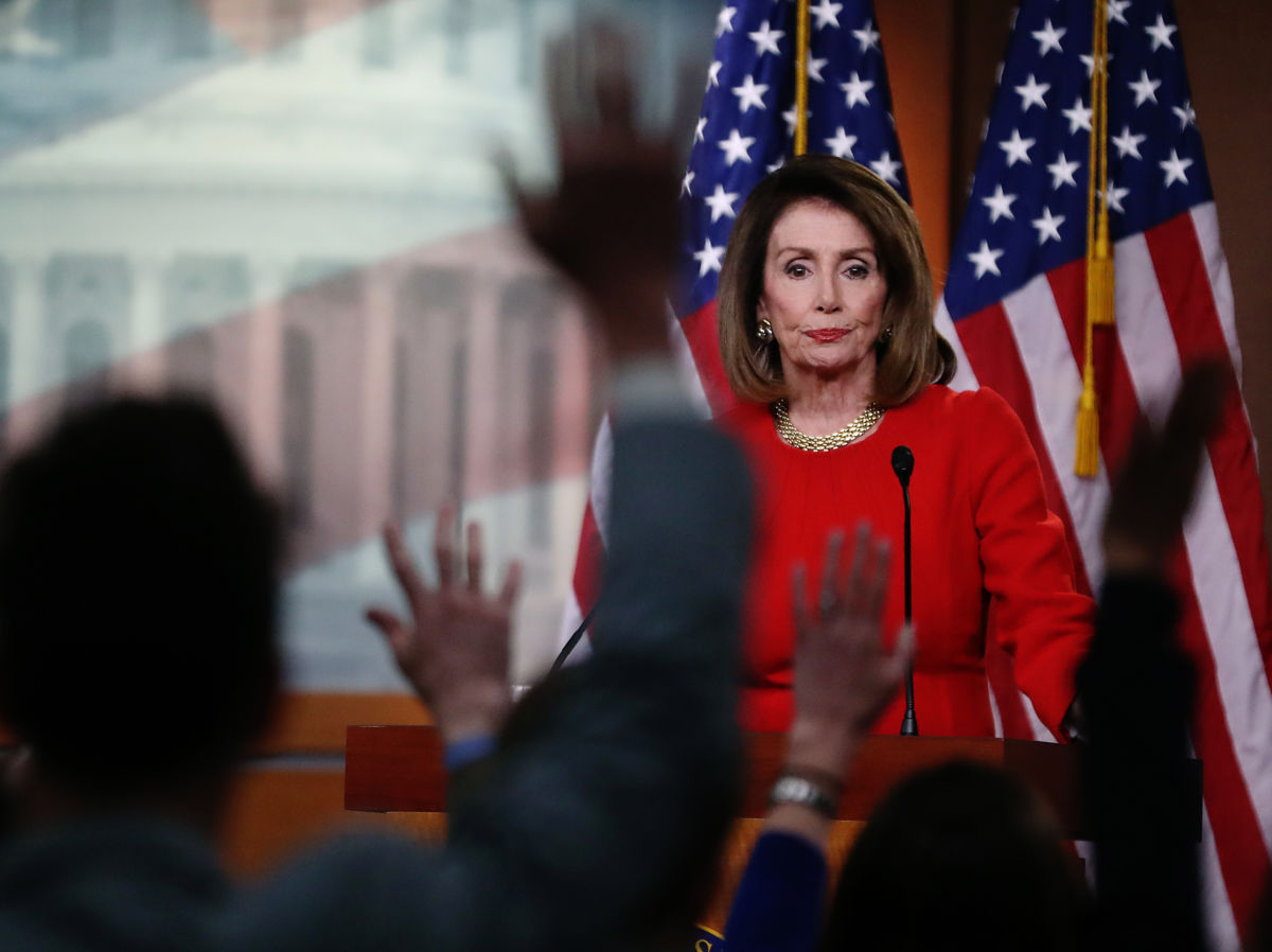 Nanci Pelosi looks on to a crowd of seated reporters raising their hands