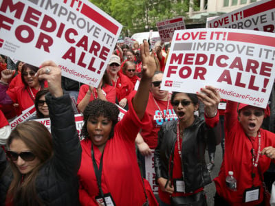 People dressed in red protest while holding "Medicare for all" signs