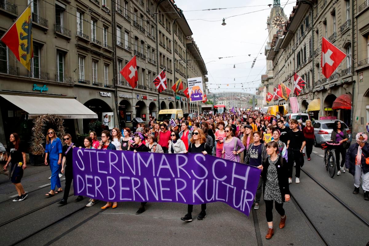 Women march through the streets holding a purple sign with a Swiss message