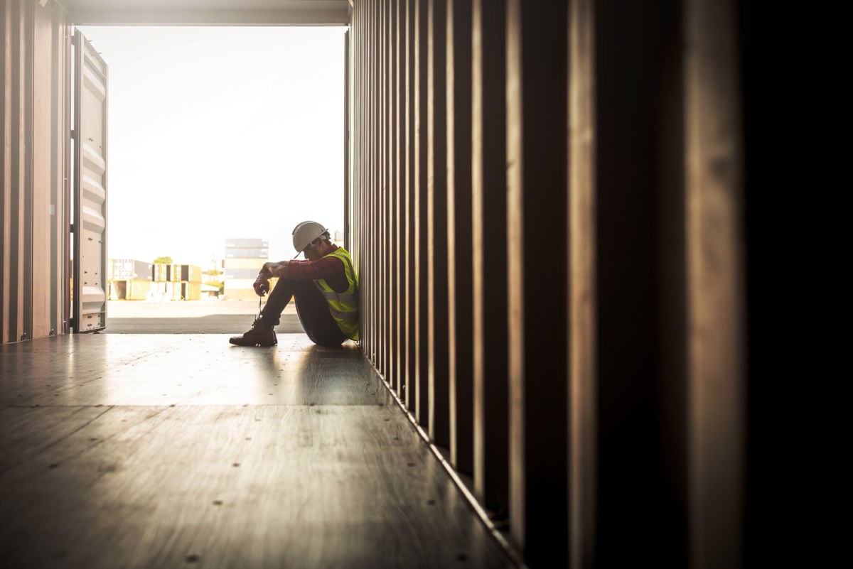 A dejected factory worker sits on the ground in an empty shipping container, unemployment, jobs